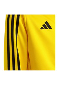 Geel sportshirt met zwarte gestreepte accenten op de schouders en een klein zwart logo met drie diagonale strepen. Te zien is een textuur van de stof.