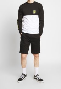 Zwart-wit shirt met lange mouwen met een geel logo, gecombineerd met zwarte korte broek en zwarte sneakers met witte veters.