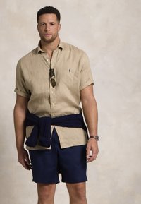Beige linnen kortärmad skjorta, marinblå shorts och en marinblå stickad tröja knuten runt midjan. Tillbehören inkluderar solglasögon och en klocka.