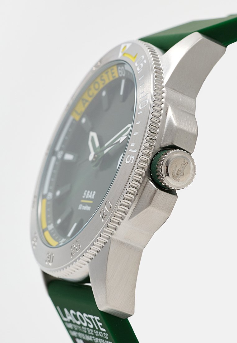 Reloj Lacoste con correa de goma verde, caja de acero inoxidable, esfera gris, manecillas plateadas, acentos amarillos y blancos, y bisel texturizado.