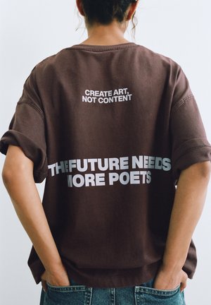 Persoană cu păr creț purtând o cămașă maro cu textul „CREATE ART, NOT CONTENT” și „THE FUTURE NEEDS MORE POETS” pe spate.