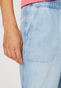 Ljusblå denimbyxor med elastisk midja, synlig sidficka och subtil textur. Handen vilar på tyget och visar designen.