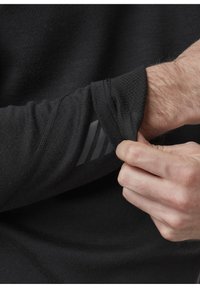 Zwarte langemouw shirt van geweven stof. Hand die de manchette aanpast, met een reflecterende gestreepte patroon op de arm.