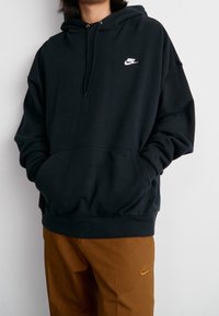 Černá mikina s kapucí s přední kapsou, stahovací kapucí a bílým logem Nike na hrudi. Měkká látka s volnějším střihem.