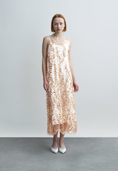 Μακρύ, αμάνικο φόρεμα σε απαλό ροζ με περίτεχνη χρυσαφένια κεντημένη διακόσμηση φύλλων, με άνετη γραμμή και τελείωμα με λεπτομέρειες δαντέλας, συνδυασμένο με λευκές ψηλοτάκουνες γόβες.