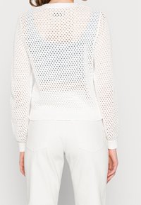 Pull en tricot léger blanc présentant un design en maille, des manches longues, des bords côtes aux poignets et à l'ourlet, ainsi qu'un dos ouvert pour la respirabilité.