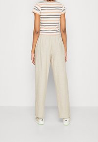 Person som står vänd bort, iförd beige vida byxor, en kortärmad randig skjorta och vita sneakers mot en enfärgad vit bakgrund.