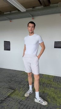 Νέος άνδρας μέσα στο σπίτι, φοράει λευκό T-shirt, ανοιχτόχρωμα ριγέ σορτς, λευκές κάλτσες και λευκά αθλητικά με μαύρες ρίγες.