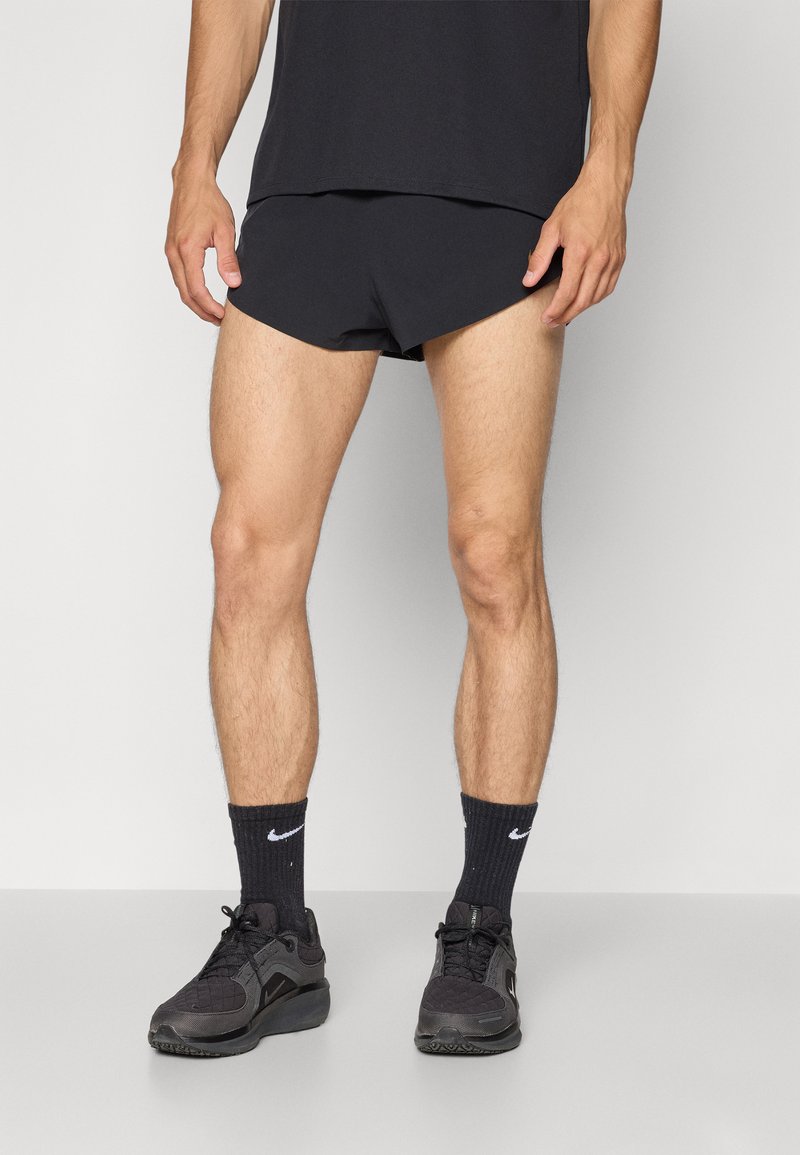 Pantalones cortos deportivos negros con un ajuste elegante, combinados con zapatillas oscuras y calcetines negros con estampado, usados con una camiseta negra. Textura de tela suave.