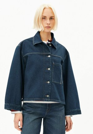 Junge Person mit kurzen blonden Haaren trägt eine dunkelblaue übergroße Jeansjacke und passende Jeans vor weißem Hintergrund.
