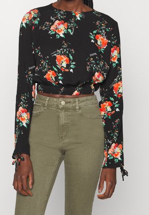 Sort langærmet cropped top med lys orange blomstermønster, kombineret med højtaljede olivengrønne bukser, båret af en stående person.