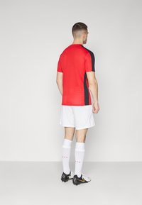 Rotes Sportshirt mit kurzen Ärmeln und schwarzen Seiten, weiße Shorts, schwarze Fußballschuhe mit gelben Akzenten und weiße Kniesocken.