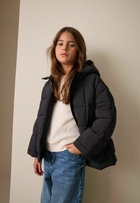 Czarna pikowana kurtka puchowa z kapturem, z bocznymi kieszeniami. Noszona na kremowej bluzie i w połączeniu z niebieskimi dżinsami.