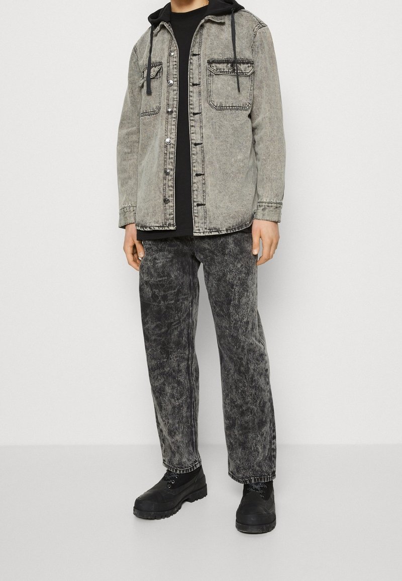 Jeune personne portant une veste en jean grise effet acide avec capuche noire, t-shirt noir, jeans gris effet acide et bottes noires à lacets, debout.