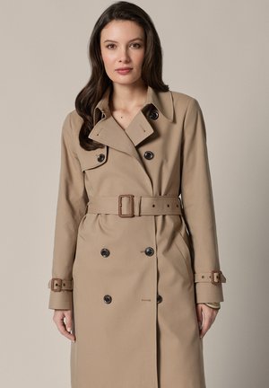 Kobieta ubrana w beżowy, dwurzędowy trencz z paskiem w talii i detalami z klamrami na mankietach na jednolitym tle.