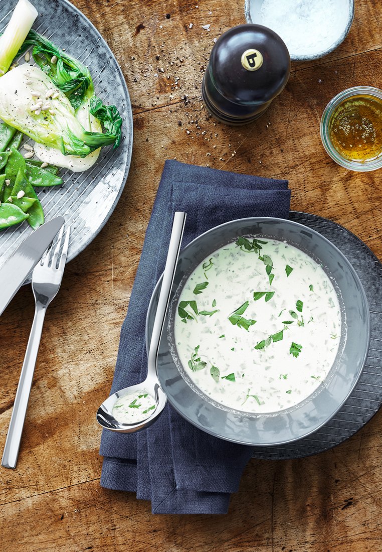 Szara miska z kremową zupą udekorowaną zielonymi ziołami, umieszczona na ciemnej, fakturowanej talerzu, na złożonym granatowym serwetniku. Srebrna łyżka i nóż.