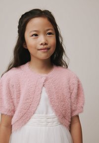 Cardigan roz pufos, cu mâneci scurte, decolteu rotund și închidere cu nasturi în față, purtat peste o rochie albă cu fustă stratificată.