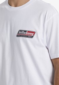 Bílé bavlněné tričko s černým a červeným logem Billabong na levé horní části hrudi, klasický kulatý výstřih a krátké rukávy.