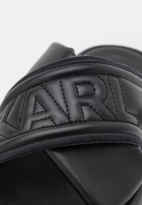 Gros plan d'une lanière de sandale en cuir noir avec couture rembourrée montrant des lettres embossées partielles "ARL".