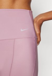Nike didelio juosmens leggingai šviesiai purpurinės spalvos, turintys lygų, tamprų audinį, plokščias siūles ir mažą šviesiai pilką logotipą šalia juosmens.