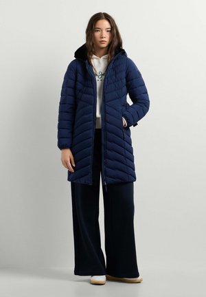 Junge Frau trägt eine lange marineblaue gesteppte Jacke über einem weißen Hoodie, weite schwarze Hose und weiße Schuhe, steht vor einem schlichten hellen Hintergrund.