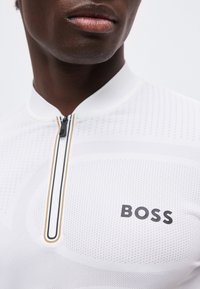 Κοντινό πλάνο ενός ατόμου που φοράει λευκό πουκάμισο με φερμουάρ, με μικρές οπές και τη λέξη «BOSS» τυπωμένη στο στήθος.
