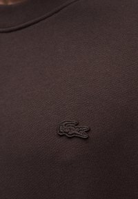 Sweat-shirt marron en tricot avec un tissu texturé et un discret logo crocodile brodé en noir sur le côté gauche de la poitrine.