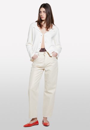 Cardigan bianco con colletto e chiusura a bottoni, abbinato a pantaloni beige chiari a gamba larga e ballerine rosse, che mostrano un fit rilassato e diverse texture.
