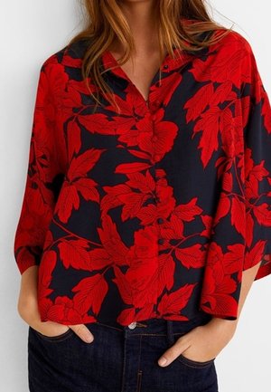 Kobieta ubrana w luźną czarną koszulę z dużymi czerwonymi kwiatowymi wzorami, ręce w kieszeniach ciemnoniebieskich jeansów, widoczna od ramion do bioder.