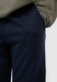 Gros plan d'une personne portant un pantalon bleu marine avec un petit logo embossé et un haut à manches longues vert olive avec un détail sur la manche.