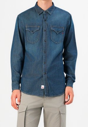 Mężczyzna ubrany w niebieską, dżinsową koszulę na guziki z długim rękawem i dwoma kieszeniami na piersi oraz jasnoszare bojówki, stojący na jednolitym tle.