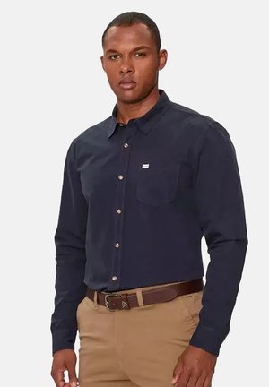 Uomo con camicia blu scuro abbottonata con taschino sul petto e pantaloni beige con cintura marrone, in piedi davanti a uno sfondo semplice.