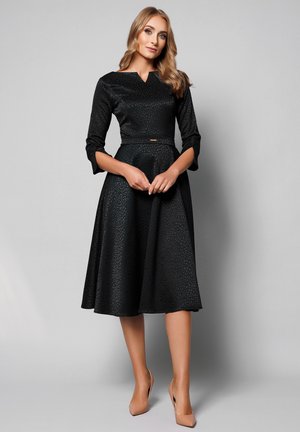 Kobieta z długimi blond włosami ubrana w czarną, teksturowaną sukienkę midi z rękawami do łokci i beżowe szpilki, stojąca na tle szarego tła.