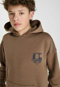 Bruine hoodie met een voorzak, voorzien van een blauw grafisch logo met "Skate & co" en "Kick & Flip." Zachte, gestructureerde stof.