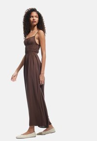 Brown sleeveless maxi dress with gathered bodice, elastic waist, flowing skirt, and soft texture, paired with light-colored flats.