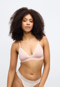 Kobieta o kręconych włosach, ubrana w jasnoróżowy biustonosz z koronkową lamówką i dopasowane koronkowe majtki, stojąca na tle jednolitego białego tła.