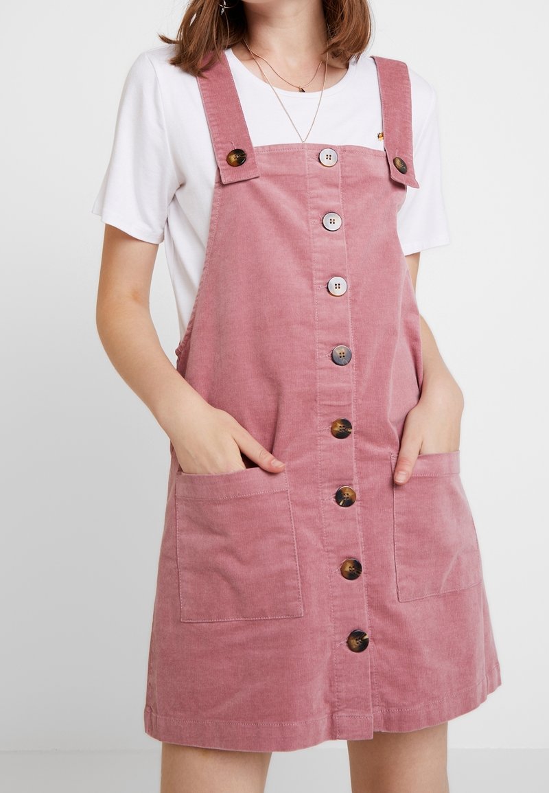 Femme portant une robe salopette en velours côtelé rose avec des boutons sur le devant et des poches à l'avant, par-dessus un t-shirt blanc à manches courtes.