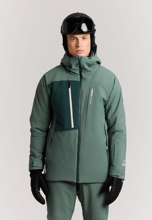 Man met groen skipak en broek, zwarte handschoenen en een zwarte skipet met skibril, die recht vooruit kijkt tegen een effen achtergrond.