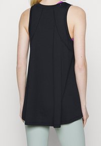 Žena oblečená v volném černém tílku bez rukávů přes purpurovou sportovní podprsenku a světle zelené legíny, zobrazená zezadu na jednoduchém pozadí.