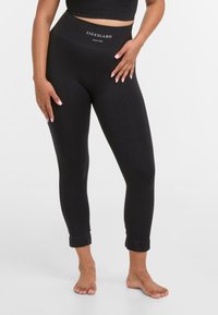 Zwarte leggings van rekbare stof, met een hoge tailleband waarop de tekst "LYKKELAND atelier" staat, een slim fit en opgerolde boorden bij de enkels.