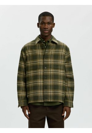 Homme portant une veste à carreaux verts et beige boutonnée, par-dessus une chemise verte et un pantalon foncé, debout devant un fond blanc uni.