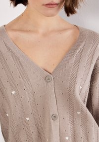 Cárdigan de punto en suave beige con escote en V, que presenta patrones texturizados, pequeños motivos de corazones y dos botones en la parte frontal.