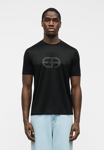 Νέος άνδρας που φοράει εφαρμοστό μαύρο t-shirt με στρογγυλό λογότυπο σε σκούρο γκρι και ανοιχτό μπλε τζιν, στέκεται μπροστά σε απλό λευκό φόντο.