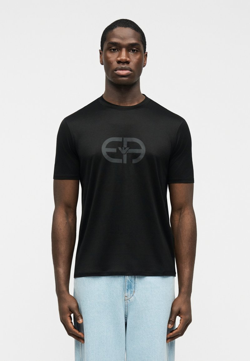 Νέος άνδρας που φοράει εφαρμοστό μ αύρο t-shirt με στρογγυλό λογότυπο σε σκούρο γκρι και ανοιχτό μπλε τζιν, στέκεται μπροστά σε απλό λευκό φόντο.