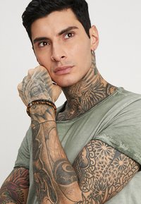 T-shirt grigio-verde, maniche corte, texture consumata. Tatuaggi visibili su braccia e collo. Braccialetto di legno al polso, orecchino a cerchio all'orecchio.