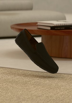 Sort slip-on loafer-sko svævende over et beige tekstureret tæppe med et rundt, træ sofabord og bøger i en sløret baggrund.