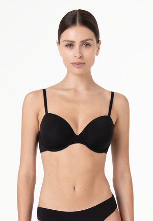 Donna con capelli scuri raccolti all'indietro che indossa un reggiseno nero e biancheria intima nera abbinata, in piedi davanti a uno sfondo bianco semplice.