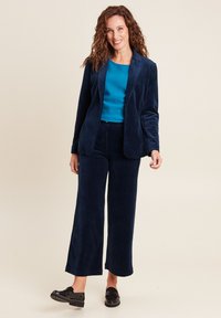 Completo in velluto a coste blu navy con blazer strutturato e pantaloni a gamba larga, abbinato a un top turchese e scarpe nere slip-on.