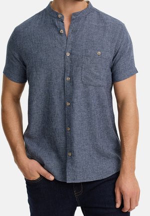 Mężczyzna noszący niebieską teksturowaną koszulę z krótkim rękawem, kołnierzykiem typu mandarynka i kieszenią na piersi, w połączeniu z ciemnymi dżinsami.