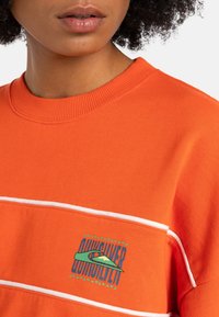 Felpa arancione con collo a girocollo a costine, caratterizzata da strisce bianche e un logo grafico blu, giallo e verde sul lato sinistro del petto.